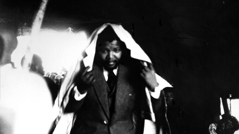 A black-and-white image of a young Nelson Mandela wearing a suit, crouching and holding a trench coat around his head, as if he is hiding himself from rain.