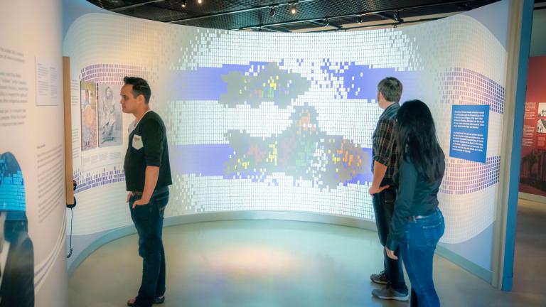 Three people stand in front of an image being projected onto a curved wall that is about nine feet tall. Two are looking at the image, while third is to the viewer’s left, at something else on the wall. The image on the wall is made up of many small squares and has the appearance of Indigenous bead work or a mosaic. The image is mainly made up of white squares, but it also features two thick blue horizontal lines that run parallel to each other. Each line is broken up near its centre by a representation of 