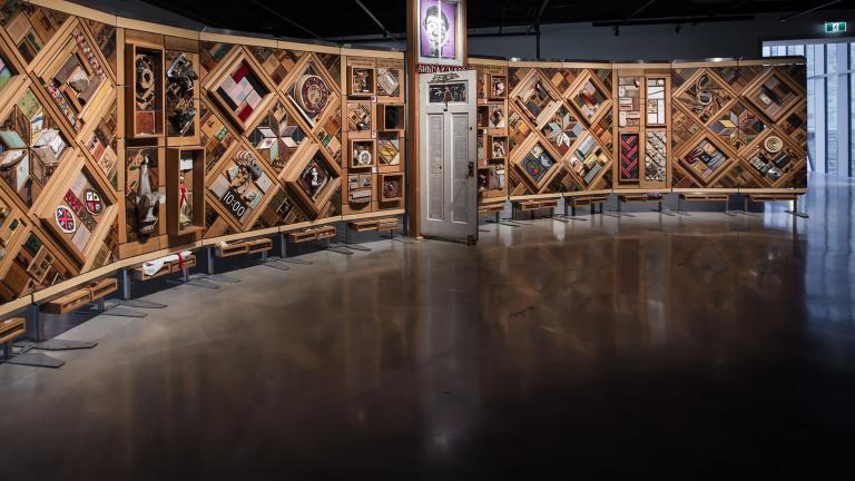 A large horizontal art installation with a wooden frame made up of multiple diamonds and rectangles to which many objects are mounted. There is a half-opened door in the middle of the work.