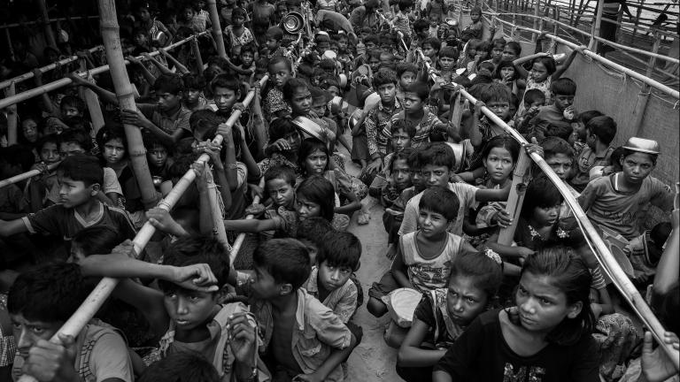 Hundreds of Rohingya children, most of them crouched or sitting down, wait in long lines for food. The line-ups are separated by long rows of bamboo poles.
