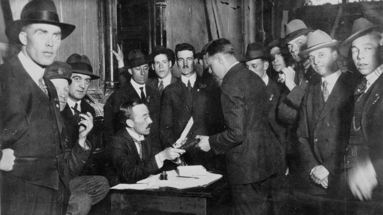 A group of men crowd around a man at a desk.