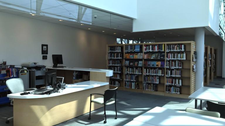 A room containing books on bookshelves, a reference desk with a computer workstation and worktables. Partially obscured.