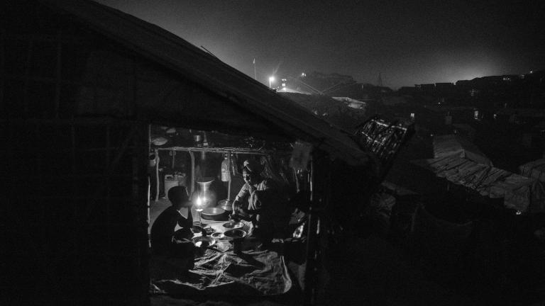 A Rohingya refugee woman serves food to a boy who sits across from her in a dark shelter. The interior is lit by a single candle.