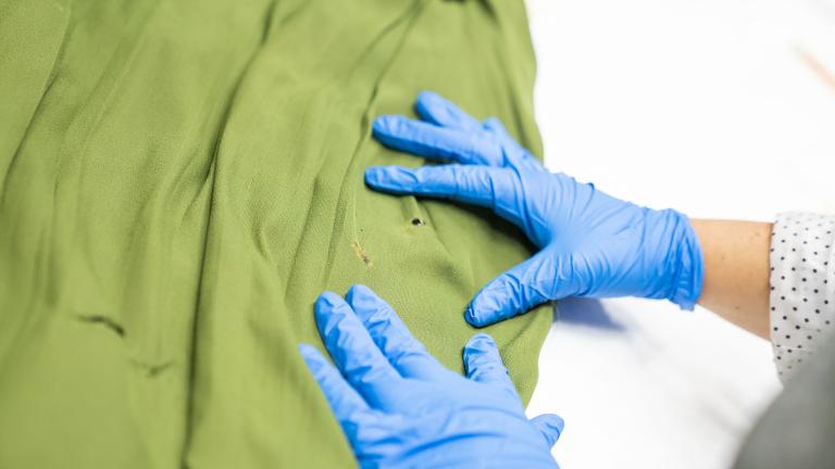 Hands with blue protective gloves are spreading apart some green fabric to show bullet holes in the fabric.