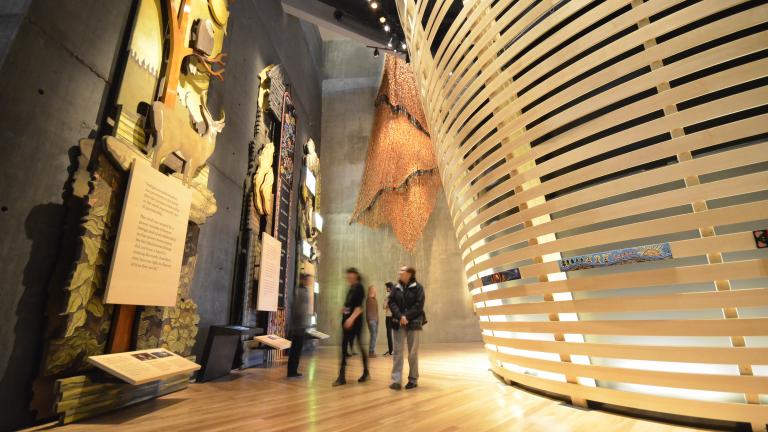 People in a museum gallery explore tall wooden panels with wildlife and Métis beadwork. There are curved horizontal wooden slats to the right.