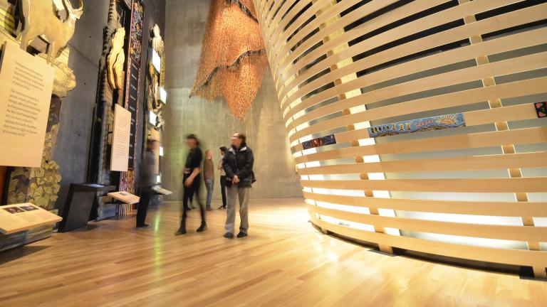 People in a museum gallery explore tall wooden panels with wildlife and Métis beadwork. There are curved horizontal wooden slats to the right. Partially obscured.