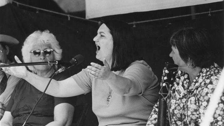 A black and white photo of a woman singing into a microphone with her arms raised. Another woman sits beside her and watches.