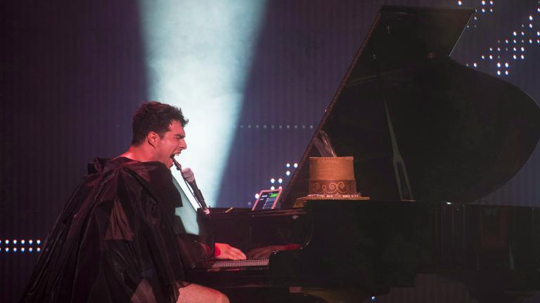 A man wearing a sheer black cape and black shorts is playing a grand piano and singing intensely into a microphone. A spotlight shines towards the ceiling behind him.