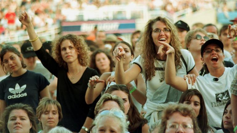 A crowd of women, viewing some sort of entertainment off-camera. Many are smiling and some have their arms raised. A larger crowd can be seen seated in a stadium behind them.