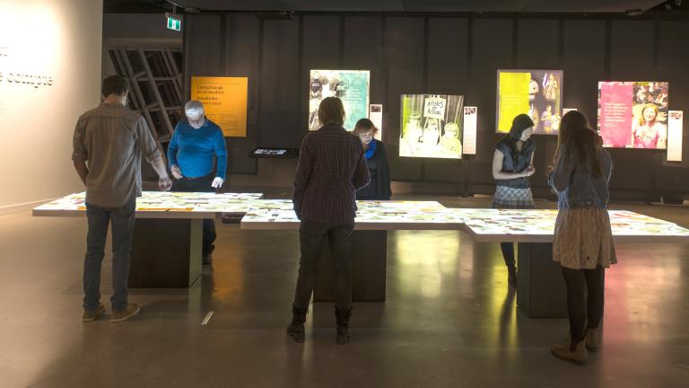 Six museum visitors observe a light table. Coloured back-lit images are on the wall.
