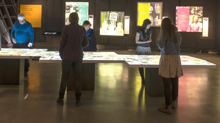 Six museum visitors observe a light table. Coloured back-lit images are on the wall.