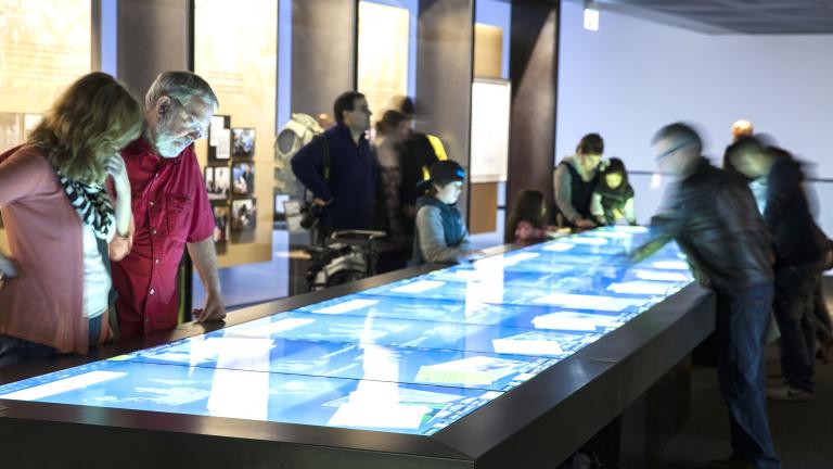 Museum visitors are looking at a large rectangular table, the surface of which is a touchscreen.