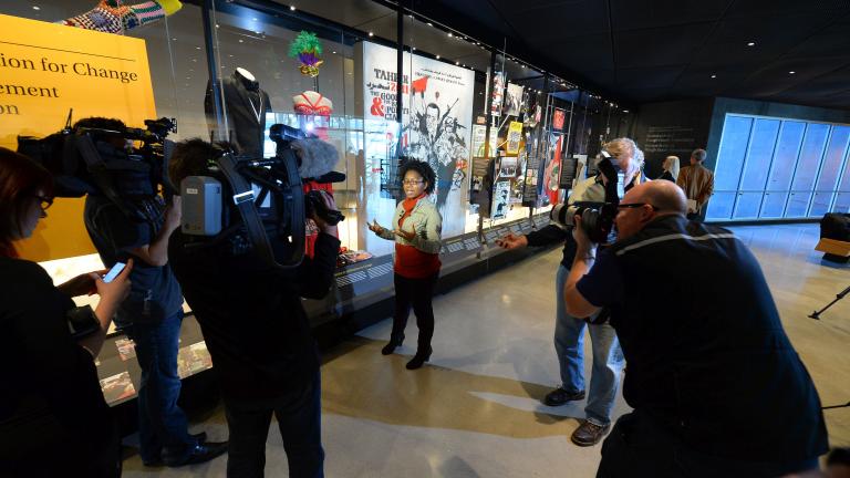 A young woman speaks while surrounded by camerapersons and photographers. Beside her is a large glass display case containing various objects, including a dress and tuxedo. 