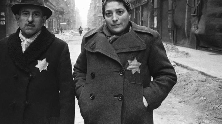 A man and a woman wearing overcoats, each with a prominent yellow star, stand in a rubble-strewn street.