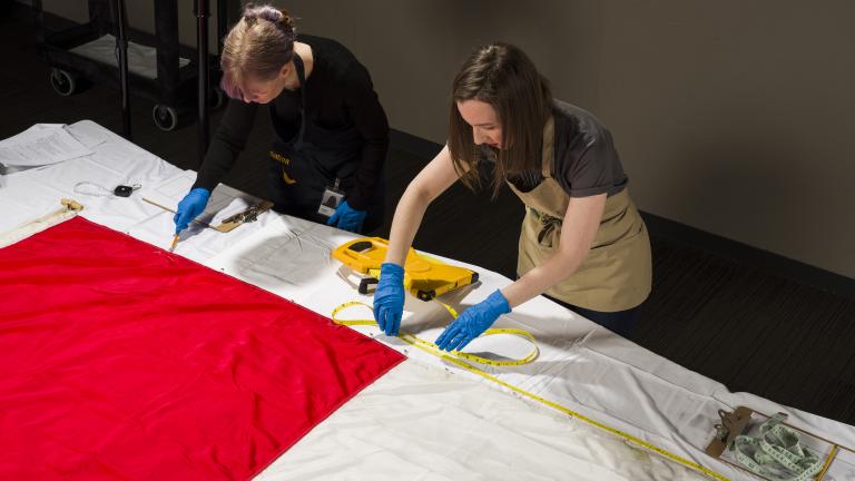 Two women wearing protective gloves and aprons measure the length of a large Canadian flag, which lies flat on a table.