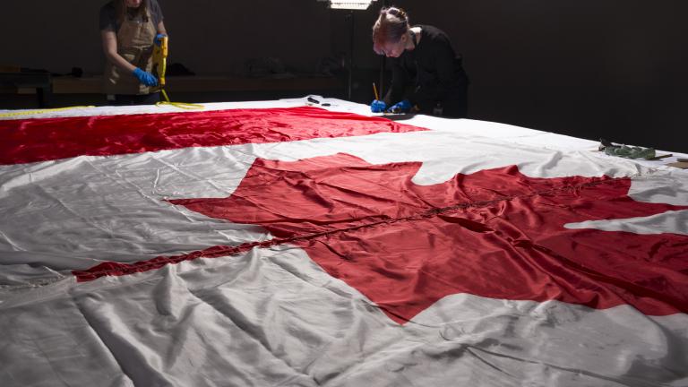 Two women wearing protective gloves and aprons working around a large Canadian flag, which lies flat on a table.