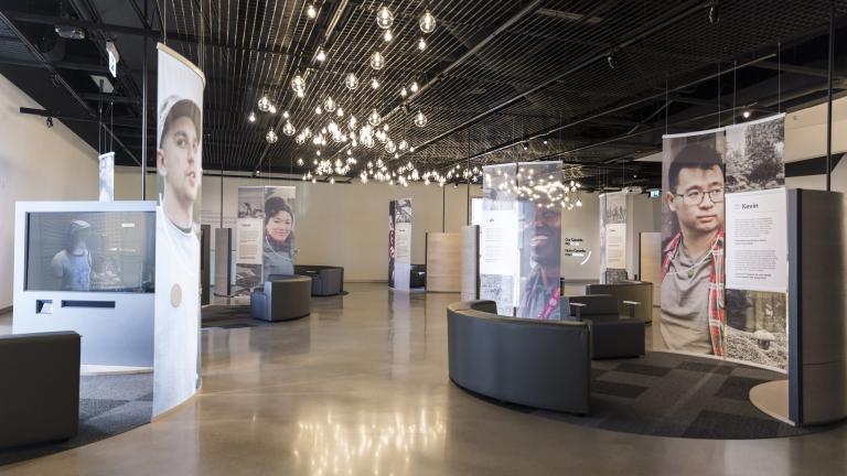 A museum display incorporating large portraits of diverse people, video screens and comfortable seating. 