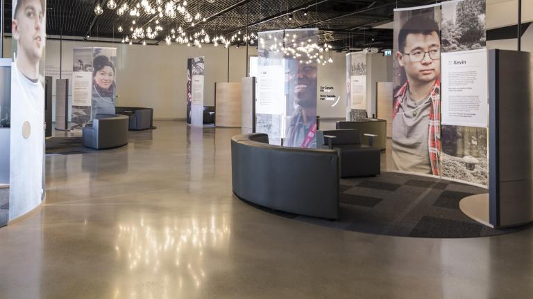 A museum display incorporating large portraits of diverse people, video screens and comfortable seating. 