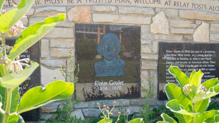 A stone wall with carved decorative script, information panels and a bas-relief portrait of a man above the words “Elzéar Goulet 1836-1870.”