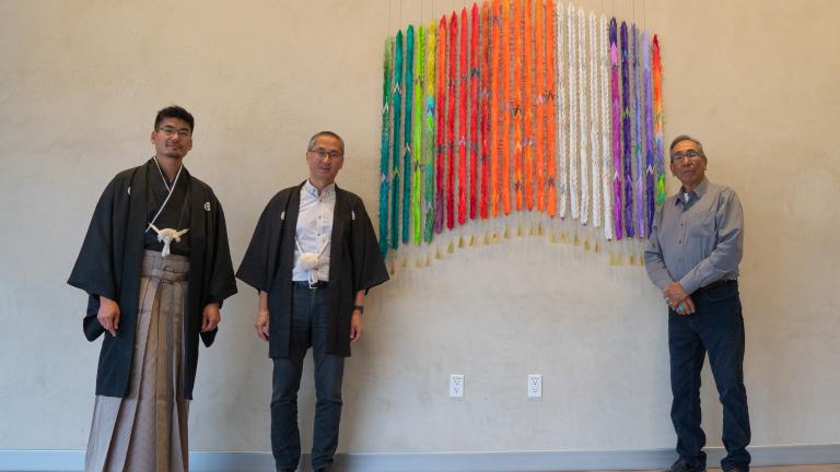 Three men stand beside a colourful artwork made of one thousand folded paper cranes. Two of the men are wearing traditional Japanese clothing. The other man is wearing a jean shirt and bolo tie.