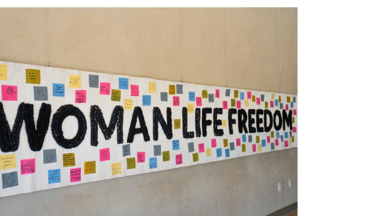 A large, white banner has “Woman, Life, Freedom” in black woven text across its centre, surrounded by 100 colourful square fabric pieces displaying solidarity messages in various languages.