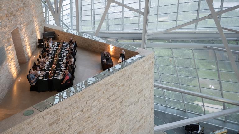 An elegant long table with people dining in an architecturally striking space, featuring tall glass windows and a high ceiling supported by metal beams.