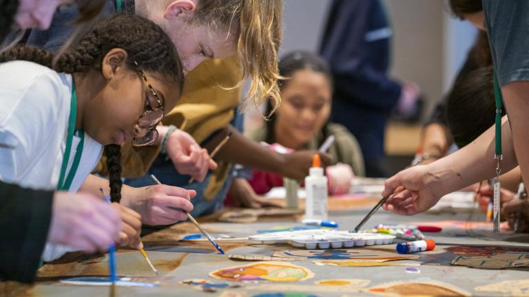 A group of children and youth paint with small brushes. They are gathered on both sides of a long table with different coloured paints in the foreground. Partially obscured.