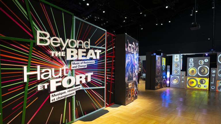 The entrance to a museum exhibit with a graphic panel includes colourful rays and text that reads “Beyond the Beat: Music of Resistance and Change” in English and French. A projected image of a pile of various speakers fills the adjacent wall. Partially obscured.