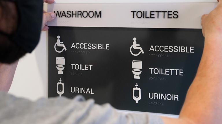 A man holds up a sign that says "Washroom" as it is mounted to a wall. Underneath the title, the words "accessible," "toilet" and "urinal" are accompanied by descriptive icons. Partially obscured.