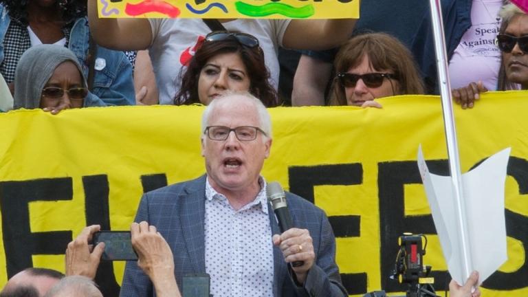 A white-haired white man with glasses and wearing a blazer is speaking and holding a microphone. Around him is a crowd of people holding cameras, protest signs and a banner. Partially obscured.