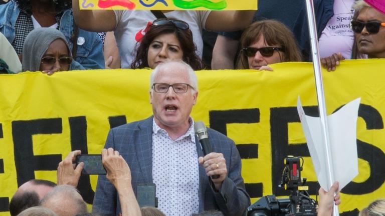 A white-haired white man with glasses and wearing a blazer is speaking and holding a microphone. Around him is a crowd of people holding cameras, protest signs and a banner. Partially obscured.