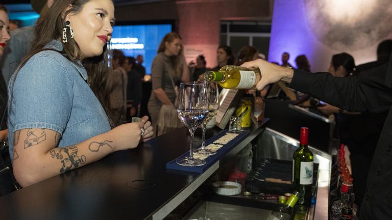 A woman waits for a drink at a bar in a museum space, while a bartender pours glasses of wine. Partially obscured.