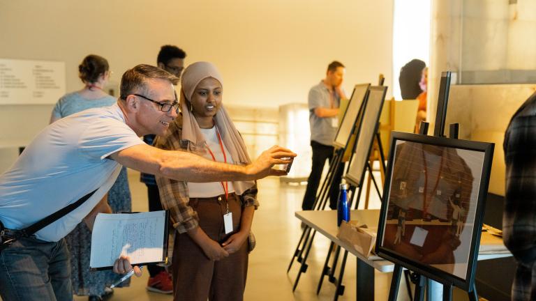Students in a museum gallery display their work on easels. The focus is on one student talking with an adult who is smiling and taking a photograph with their phone of the student’s work. The artwork shows four people in a hallway with four doors, each with a sign on it. Partially obscured.
