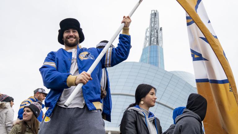 A smiling man in a crowd of people holding a large yellow flag with a blue and white, “W” in the centre. The man is wearing a blue Winnipeg Blue Bombers jacket as he stands in front of the Canadian Museum for Human Rights. Partially obscured.