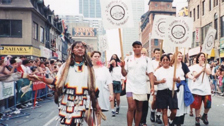 Un groupe de personnes portant des pancartes sur lesquelles on peut lire « 2-Spirited People of the 1st Nations » marchent au centre d’une rue de la ville, tandis que des gens regardent derrière des barrières de part et d’autre de la rue. Partially obscured.