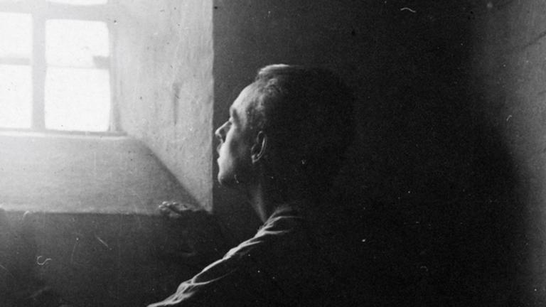 A man stands in dark shadows in a stark stone cell, gazing upward at bright sunlight streaming in through a barred window. Partially obscured.