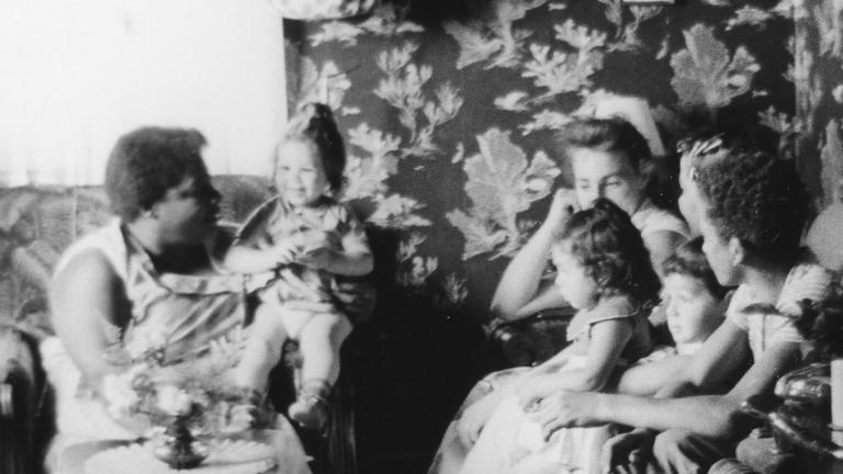 A black and white image of women and young children sitting on couches and chairs in a small living room.