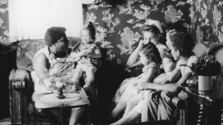 A black and white image of women and young children sitting on couches and chairs in a small living room. Partially obscured.