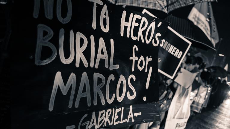 People standing in the rain under umbrellas along a roadside. Headlights from vehicles passing them are on the right side of the image. In the left foreground a sign blocks their faces from view and reads “No to hero’s burial for Marcos! Gabriela.” Partially obscured.