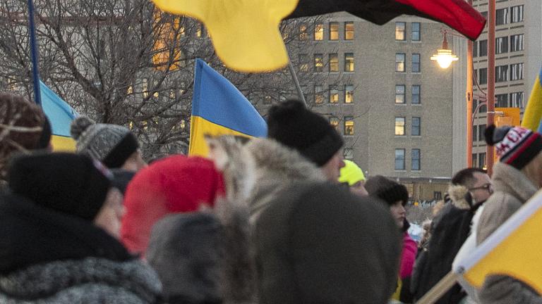 Une foule de personnes emmitouflées se tient debout dans une rue de ville pendant l’hiver, tenant de multiples drapeaux ukrainiens bleus et jaunes ainsi qu’un drapeau canadien. Le rassemblement semble être une manifestation en appui à l’Ukraine. De grands édifices à bureaux et des feux de circulation sont à l’arrière-plan sous un ciel couvert. Partially obscured.