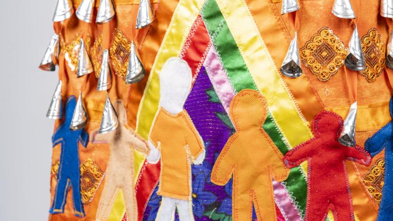 A section of an orange jingle dress is shown. Jingles, or small silver cone-shaped pendants, are visible at the top of the image. Under them, seven human figures are shown, all in various colours and sizes. One of the figures is wearing an orange shirt. These human figures are holding hands inside a multicoloured tipi. Partially obscured.