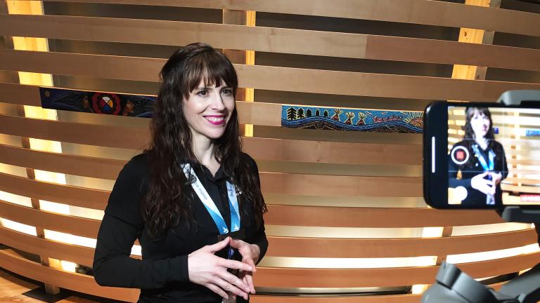 A smiling woman with light skin is filmed on a cell phone. She is wearing a lanyard with the words “Be inspired” and standing in front of an exhibit element made of horizontal strips of wood, some decorated with Indigenous art. Partially obscured.