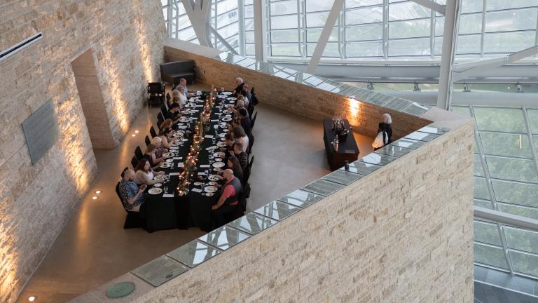 An elegant long table with people dining in an architecturally striking space, featuring tall glass windows and a high ceiling supported by metal beams. Partially obscured.