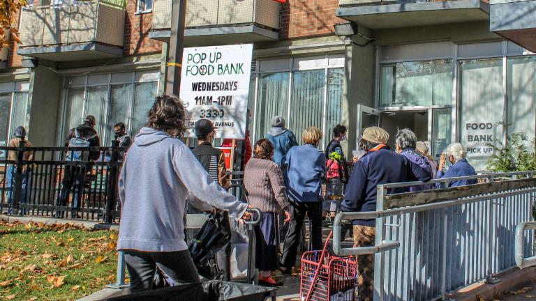 A line-up of people wait outside a building. There are signs indicating it is a Food Bank. A Canadian flag flies above them. Partially obscured.