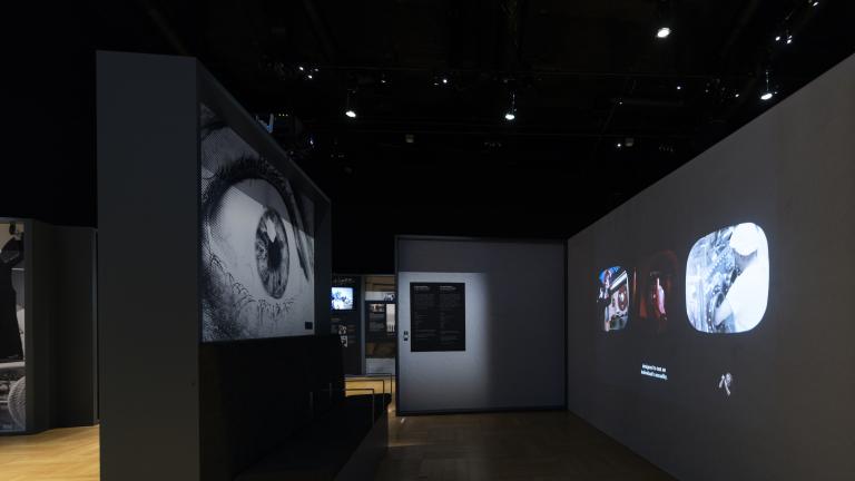 An exhibition panel on the left has a picture of a large eye on it. On the right, three small videos are projected on to the wall. The space is dark.