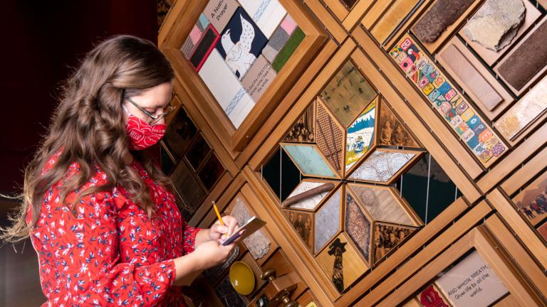 A woman wearing a facemask writes in a notepad while standing in front of a large, wood-framed art installation that is full of objects.