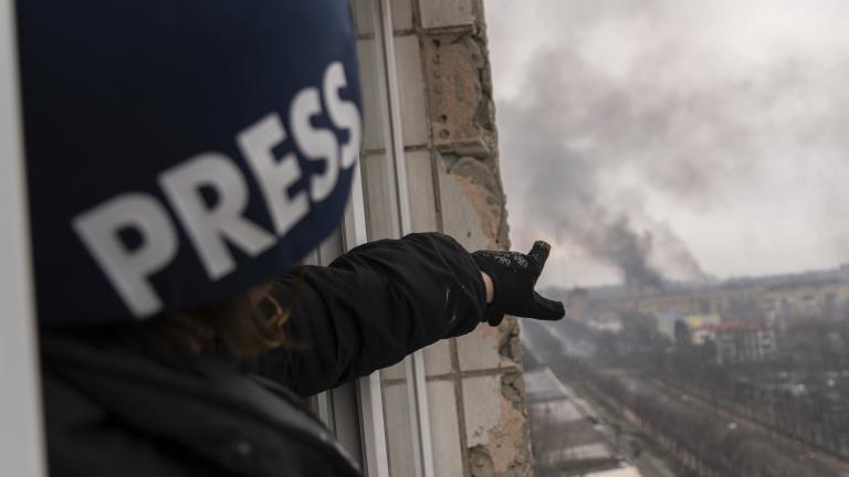 A person wearing a dark blue helmet marked with the word “PRESS” points from a balcony window high above a road, towards a pillar of smoke following an explosion. Partially obscured.