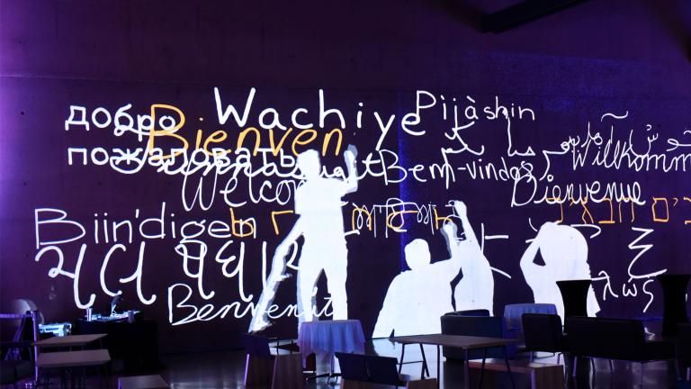 Still image from an animation showing human figures writing “welcome” in multiple languages in white and yellow on a purple wall. Partially obscured.