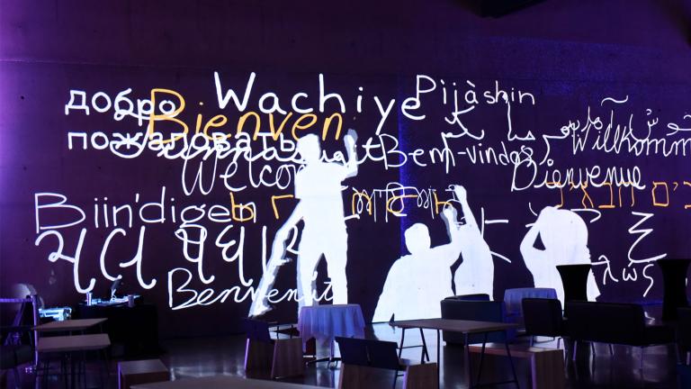 Still image from an animation showing human figures writing “welcome” in multiple languages in white and yellow on a purple wall. Partially obscured.