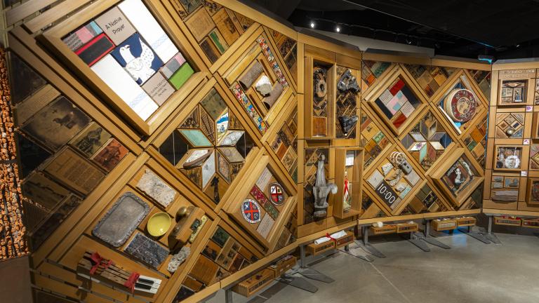 A large, curved freestanding wooden wall on which is mounted numerous small objects arranged in diamond and rectangular patterns. There is an open door in the centre of the wall. Partially obscured.